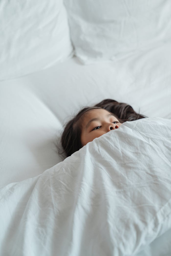 A child peacefully lying in bed, covered with a white blanket, creating a cozy and relaxing morning scene.
